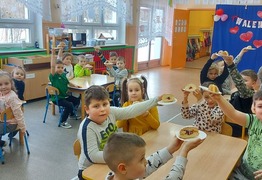 Dzieci z grupy Marzyciele (Borówko Stare) podczas świętowania dnia Tłusty Czwartek. Dzieci siedzą przy stolikach i trzymają uniesione w górze talerzyki z pączkami (photo)
