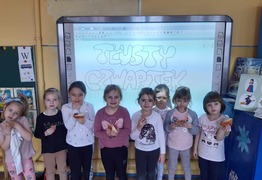 Dzieci z grupy Zajączki (Jarogniewice) podczas świętowania dnia Tłusty Czwartek. Dzieci stoją z pączkami w rękach, w tle napis „Tłusty Czwartek” (photo)