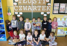 Dzieci z grupy Misie (Jarogniewice) podczas świętowania dnia Tłusty Czwartek. Dzieci stoją talerzami, na których znajdują się pączki, w tle napis „Tłusty Czwartek” i sylwety pączków (photo)